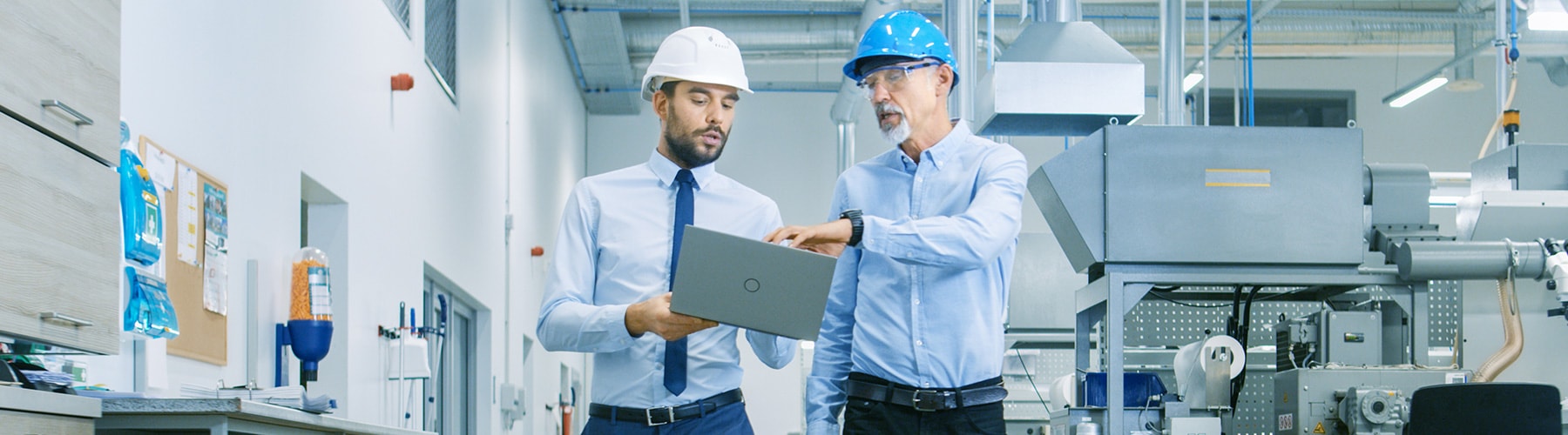 an engineer in a factory holding a laptop, in conversation with a colleague